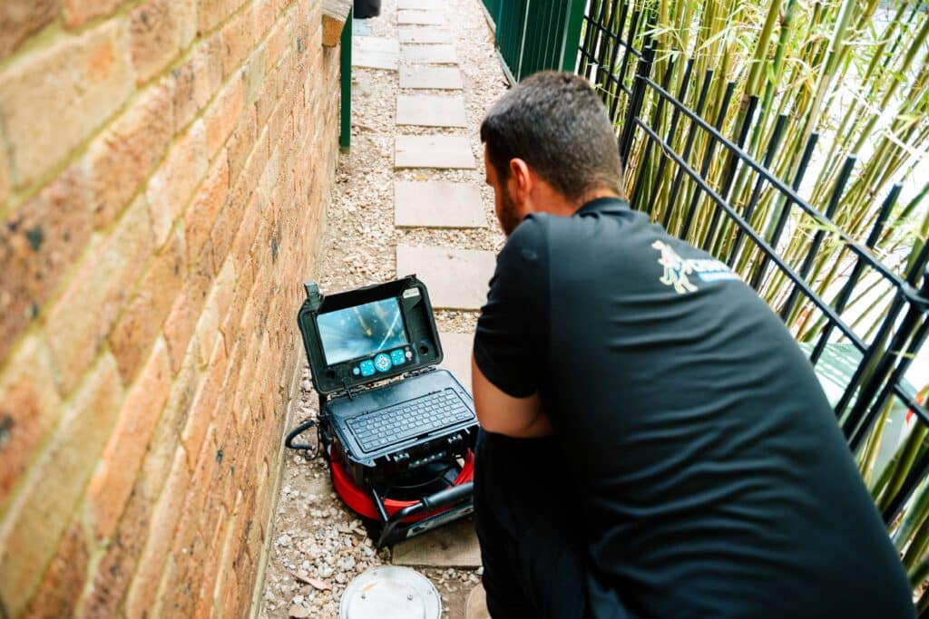 Champion Plumbing and Drains technician monitoring a live CCTV drain inspection on a screen in a narrow side passage at a residential property