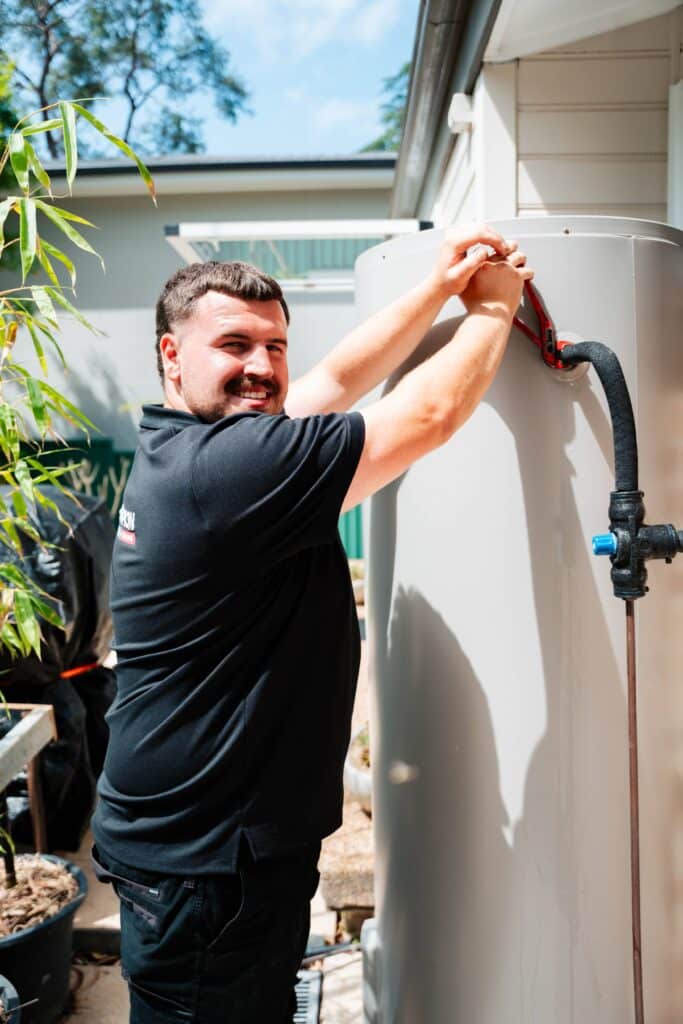 Champion Plumbing and Drains technician smiling while servicing an external hot water unit at a residential property