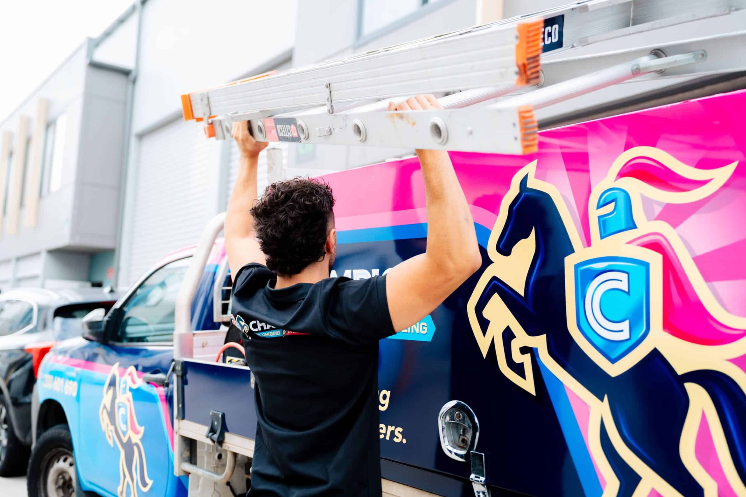 Champion Plumbing and Drains technician lifting a ladder onto the roof rack of a vivid pink branded service truck displaying the company's knight mascot