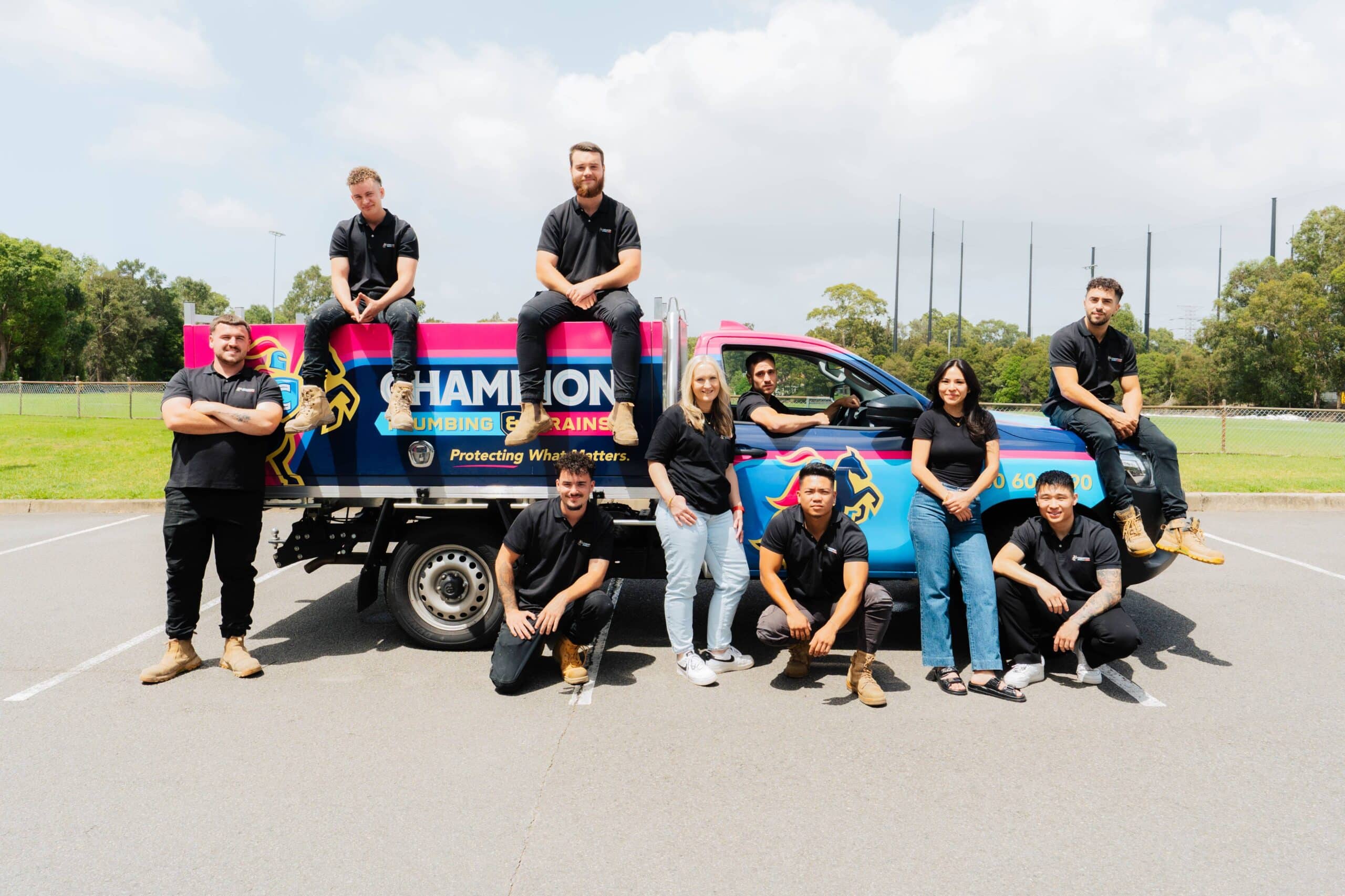 Champion Plumbing and Drains team posing in a creative group shot around two branded vehicles, with members sitting on the trucks and others kneeling and standing in front