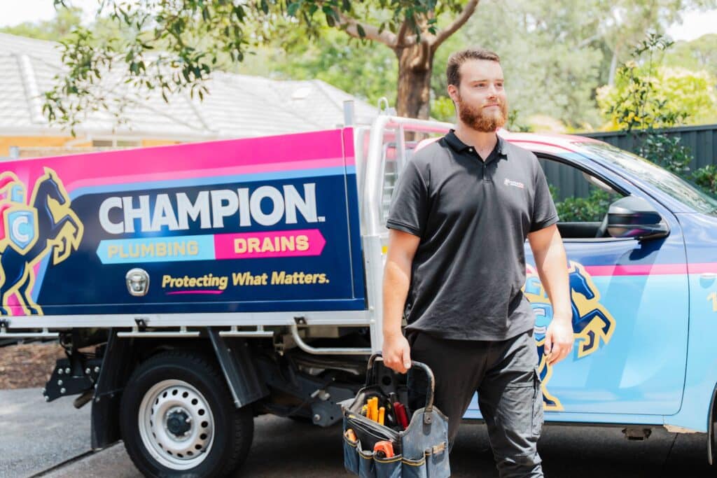 Champion Plumbing and Drains technician carrying a tool bag beside a branded service truck at a residential job site, with the Plumbing and Drains tagline clearly visible