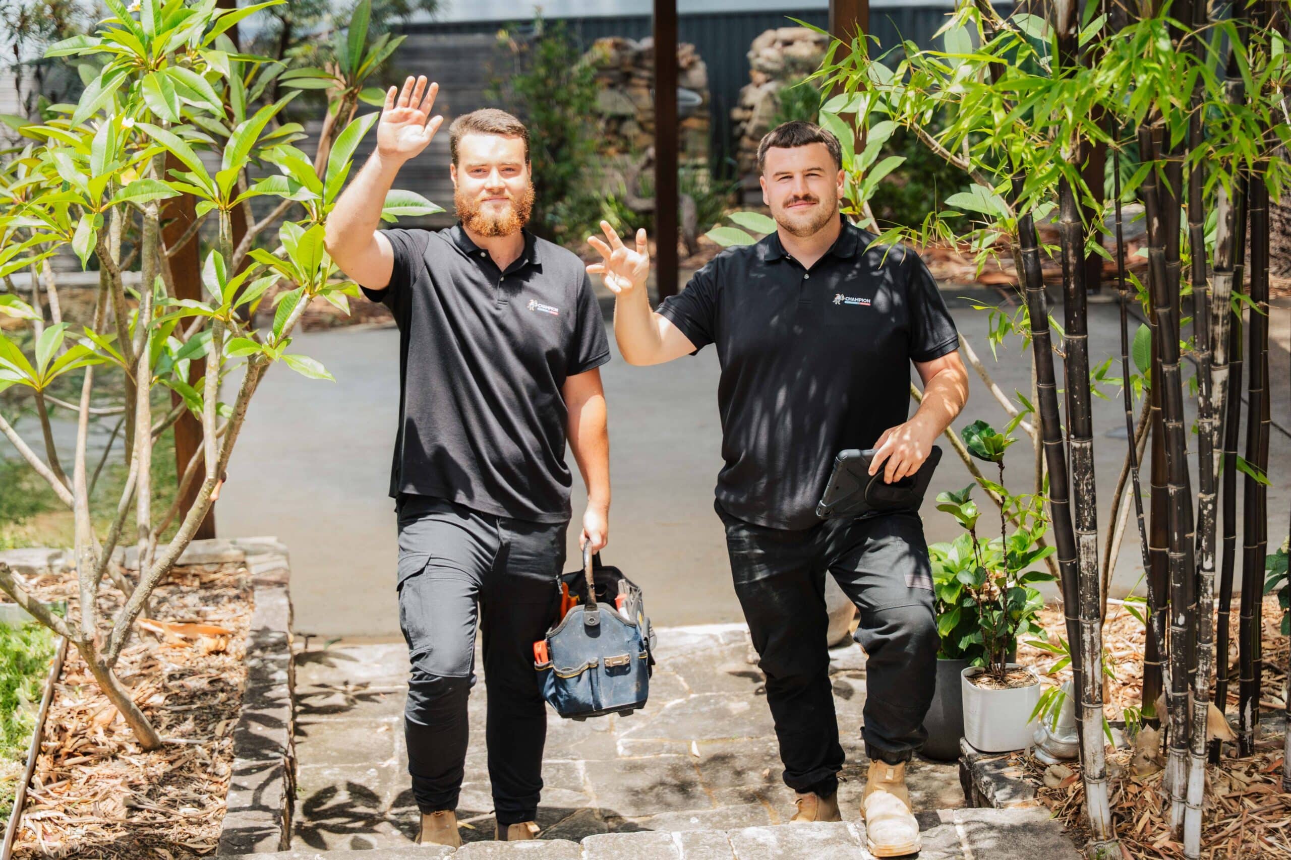 Two Champion Plumbing and Drains technicians walking through a residential garden and waving as they arrive at a customer's home
