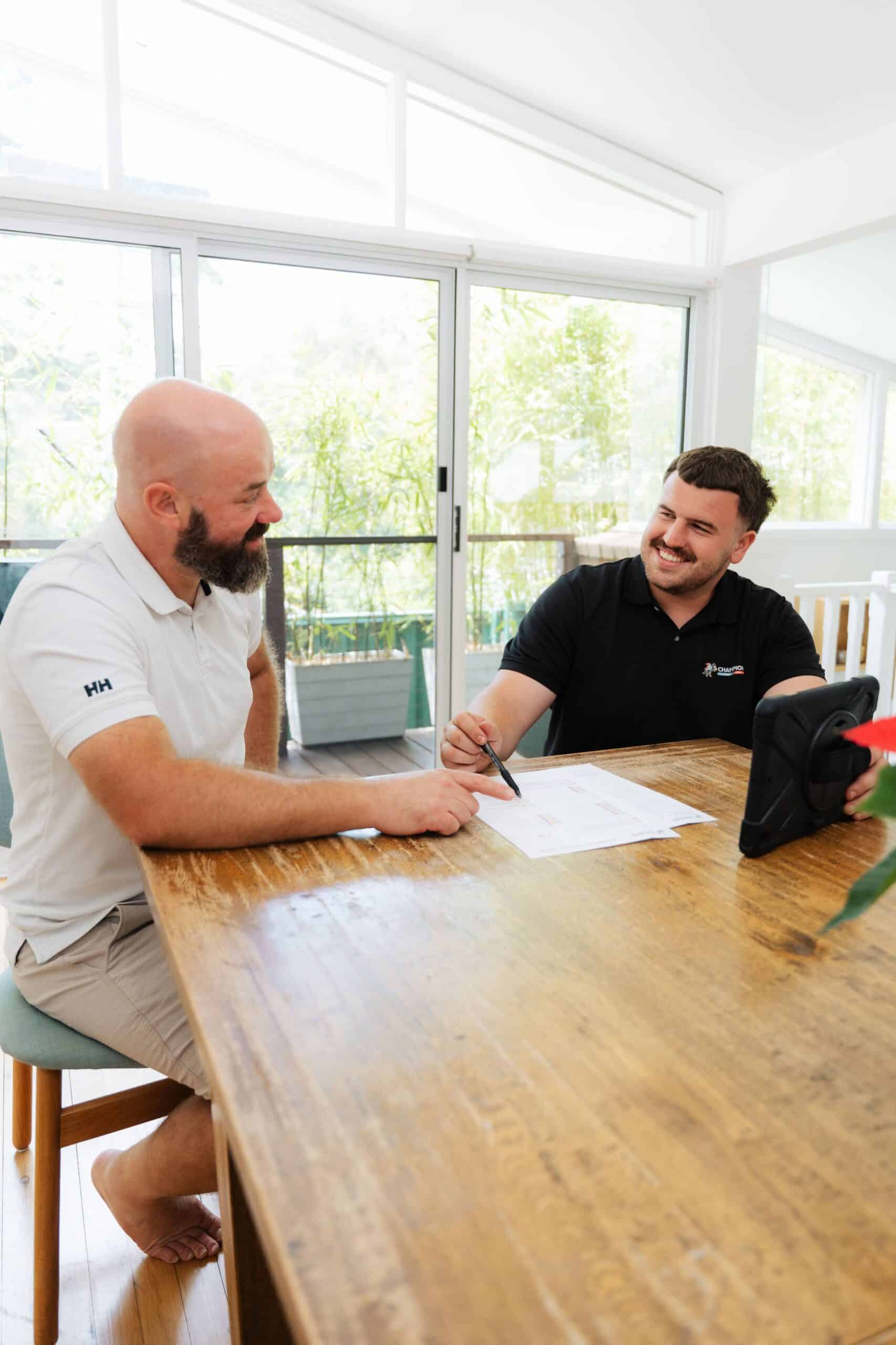 Champion Plumbing and Drains technician smiling while going over a job quote with a customer at a bright, sunlit dining table