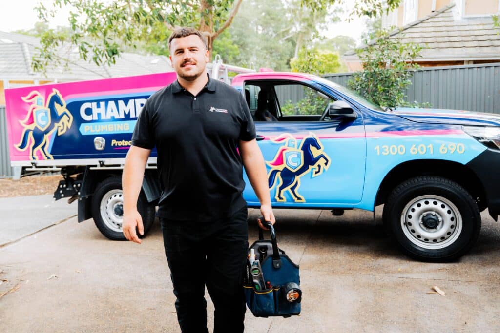 Champion Plumbing and Drains technician carrying a tool bag beside a branded service truck parked in a residential driveway