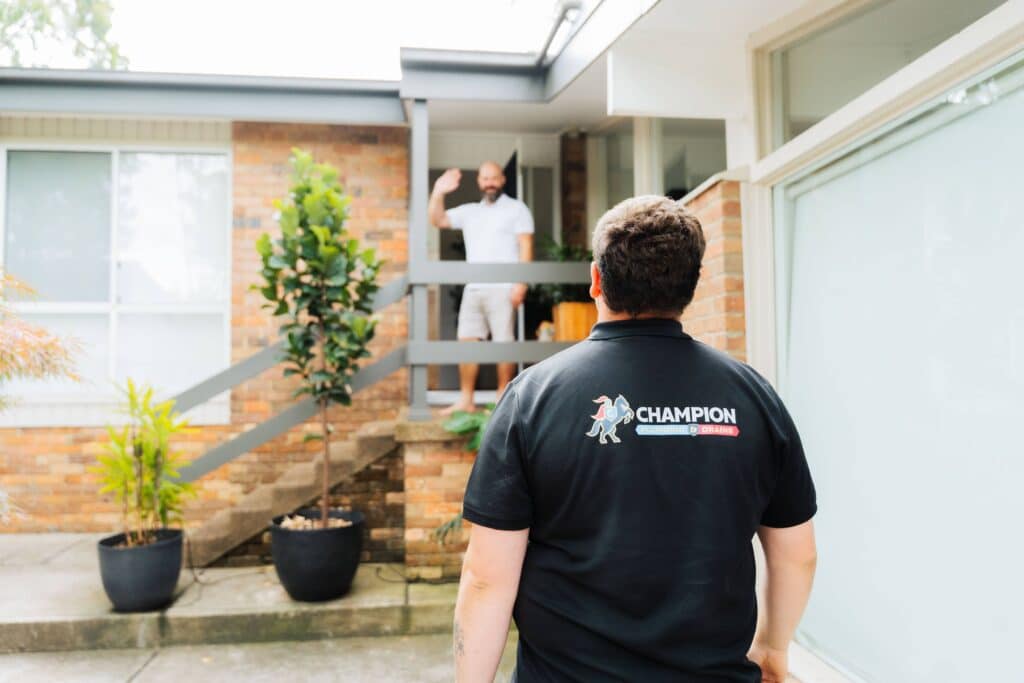 Champion Plumbing and Drains technician approaching a residential home as the customer waves from the front doorway, with the company logo visible on the back of the uniform