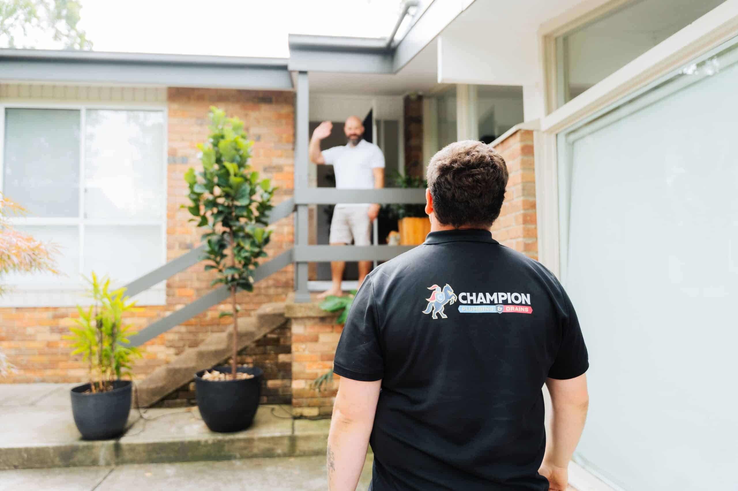 Champion Plumbing and Drains technician approaching a residential home as the customer waves from the front doorway, with the company logo visible on the back of the uniform
