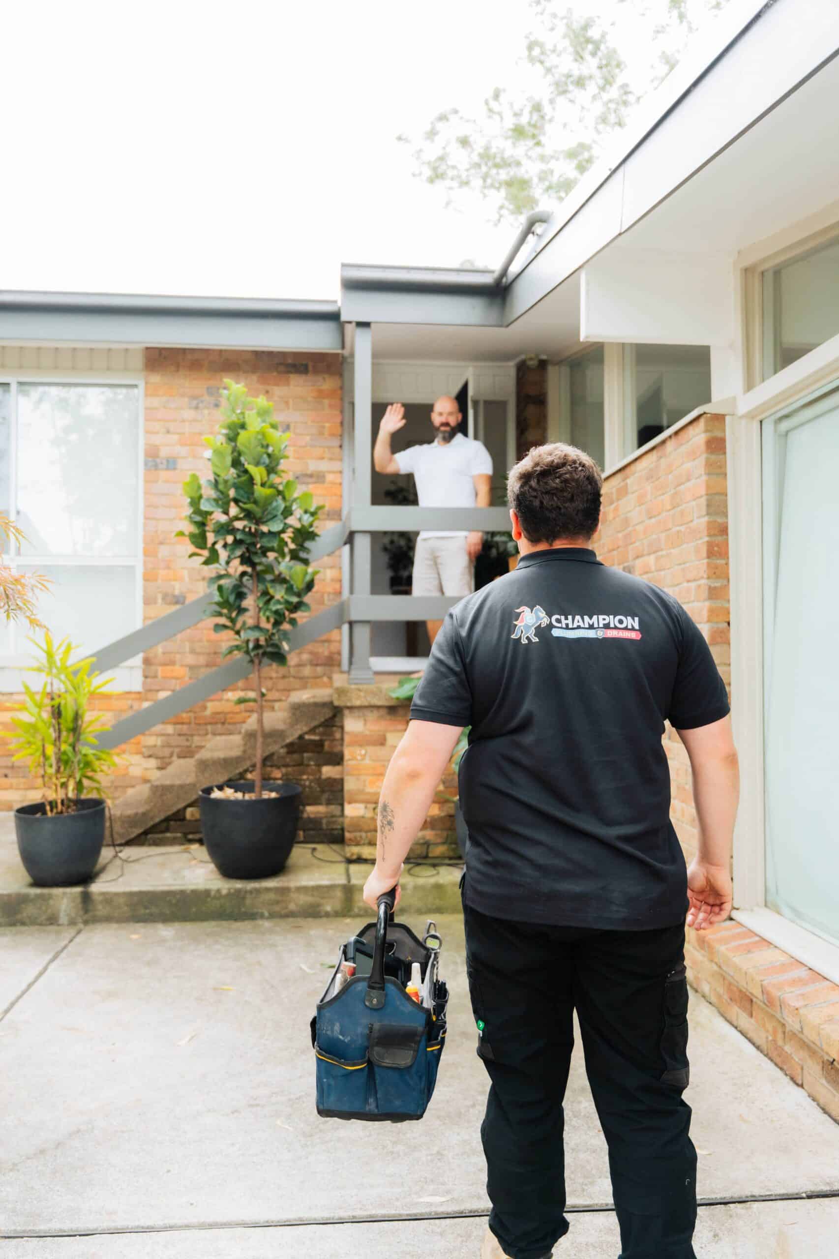 Champion Plumbing and Drains technician carrying a tool bag as he approaches a residential home where the customer is waving from the front door