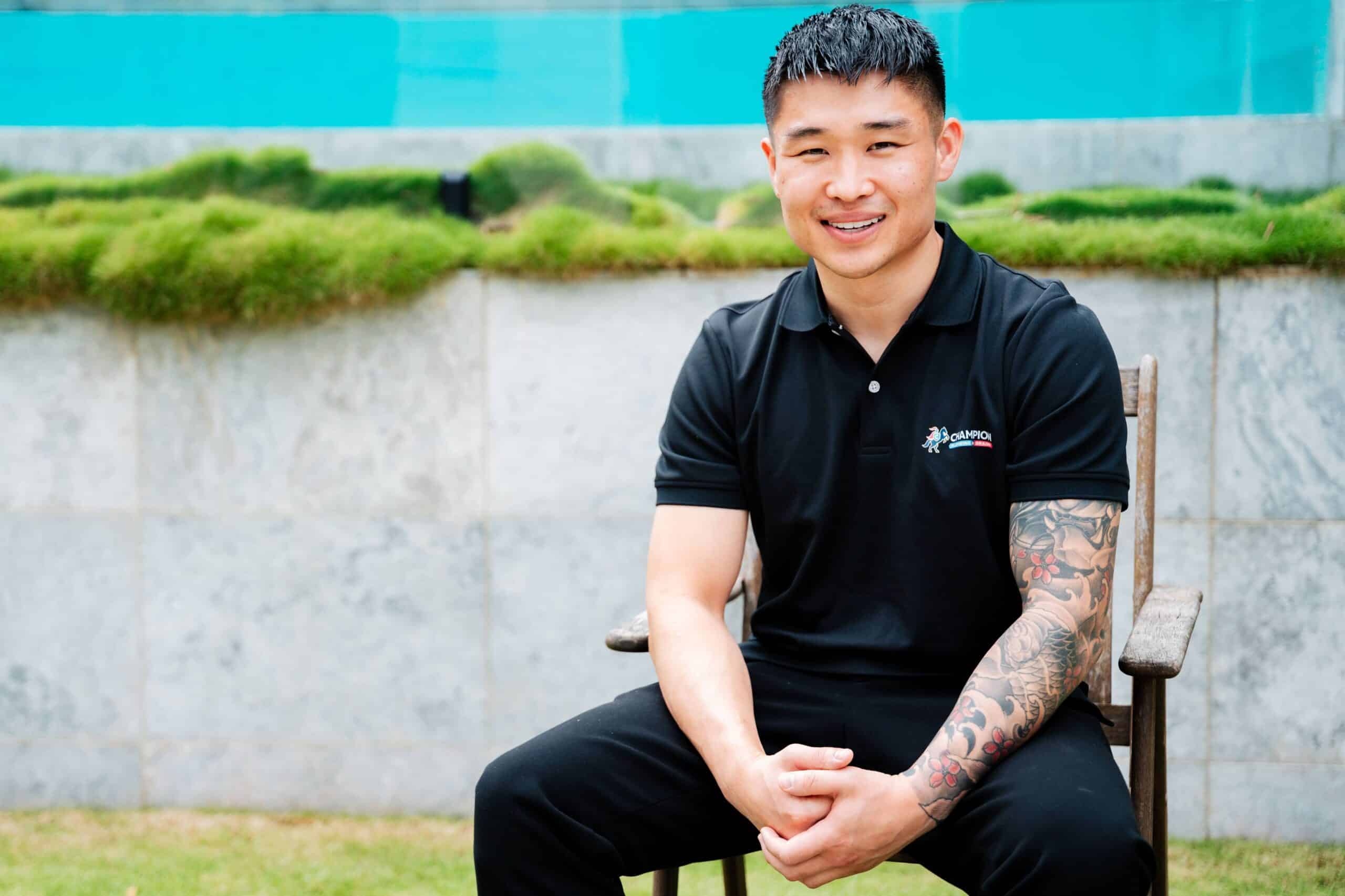Champion Plumbing and Drains founder smiling in a seated outdoor portrait with a pool and stone wall backdrop