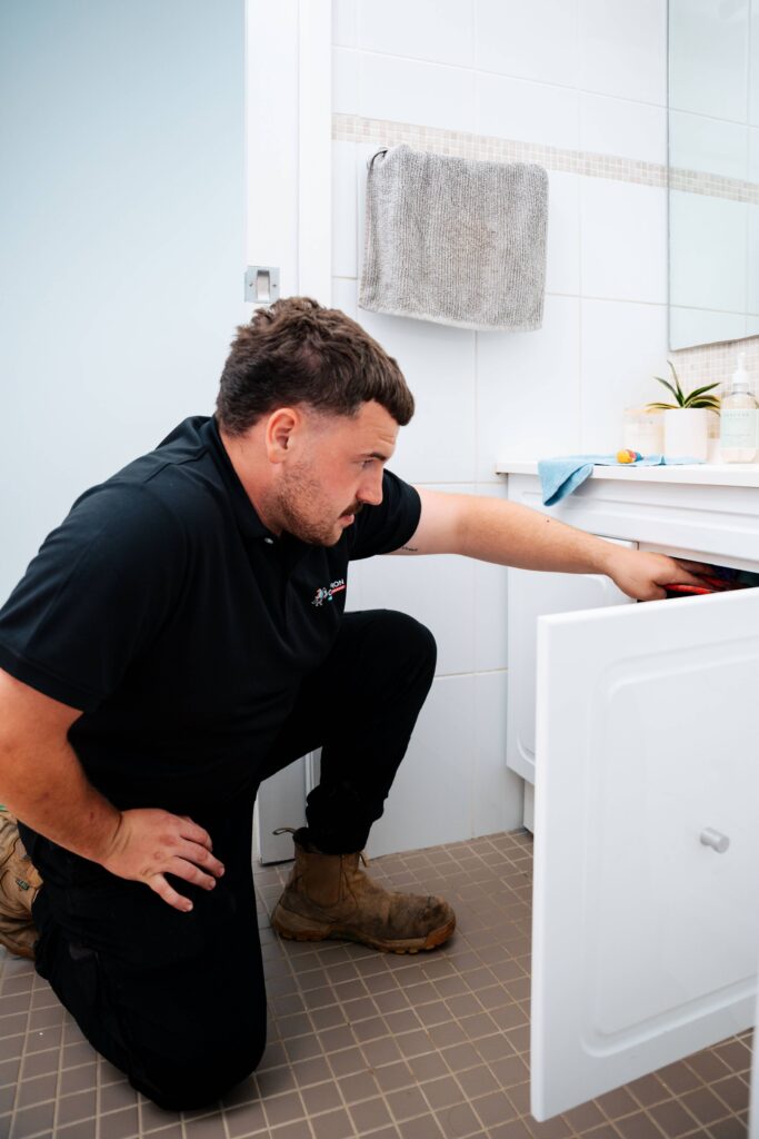 Champion Plumbing and Drains technician crouching to inspect the underside of a bathroom vanity cabinet