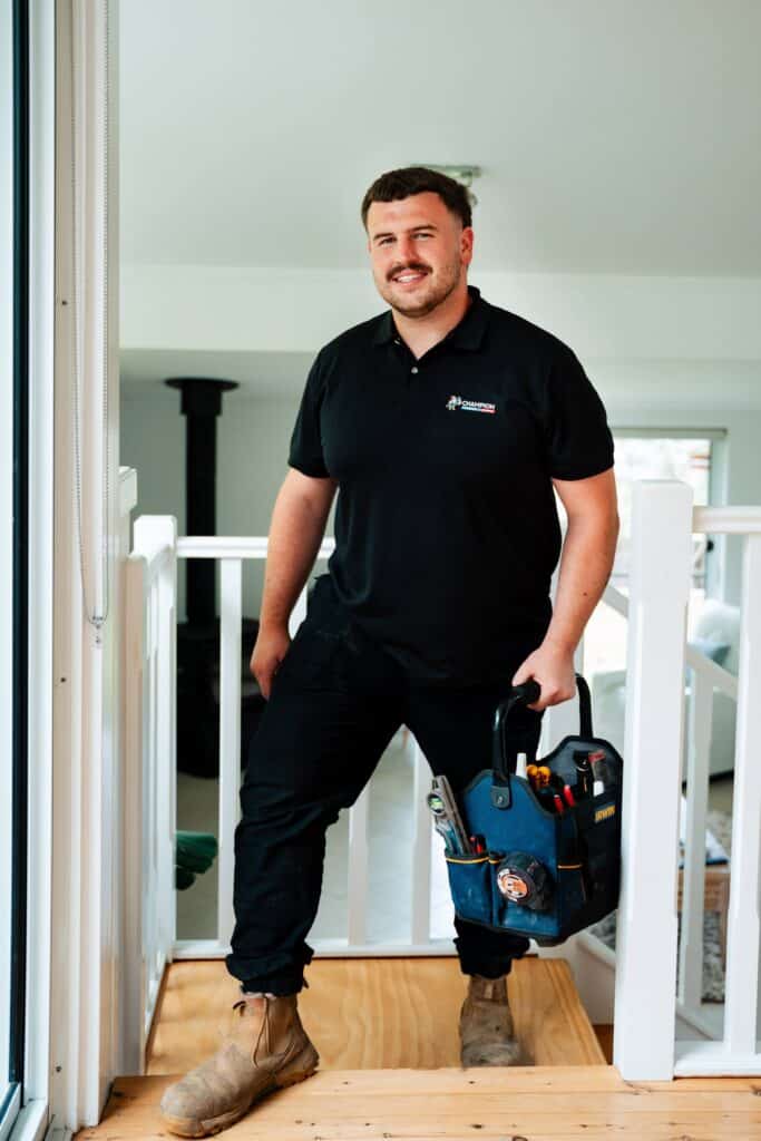 Champion Plumbing and Drains technician carrying a tool bag inside a residential home, smiling on the landing of a staircase