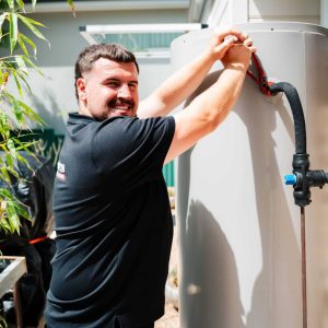 Champion Plumbing and Drains technician smiling while servicing an external hot water unit at a residential property